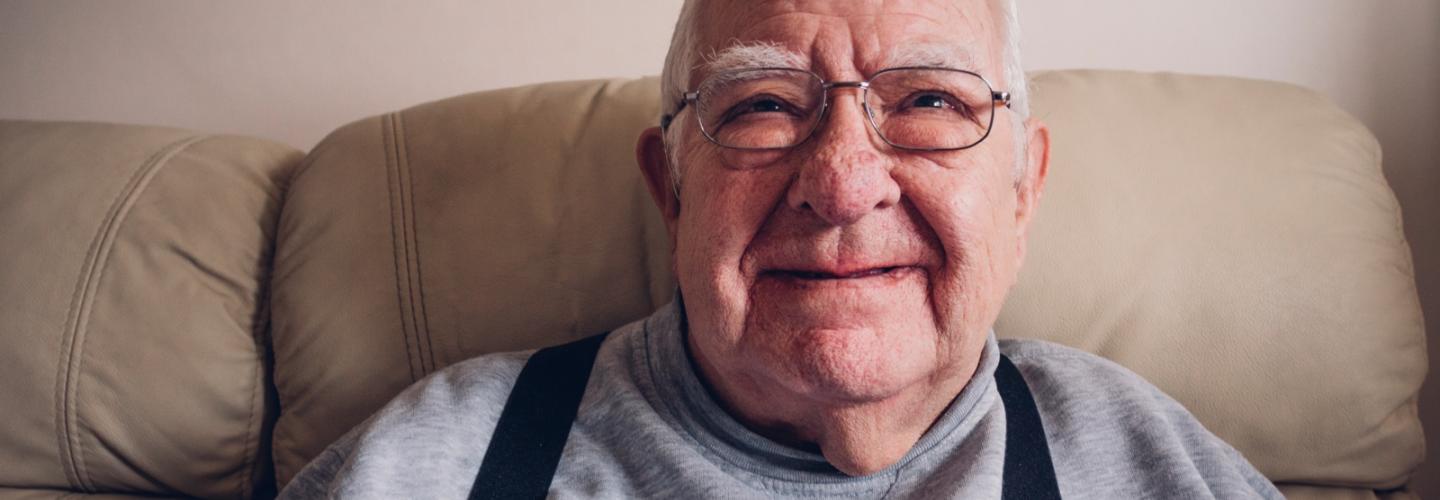 Older gentleman with glasses on sitting on a sofa smiling 