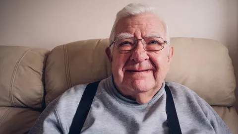 Older gentleman with glasses on sitting on a sofa smiling 